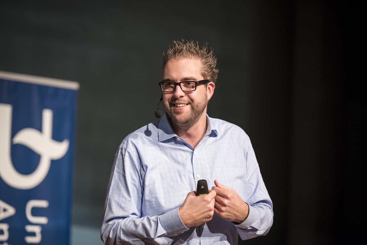 A speaker in a light blue shirt gives a presentation at Antwerp University, holding a remote and wearing a headset.