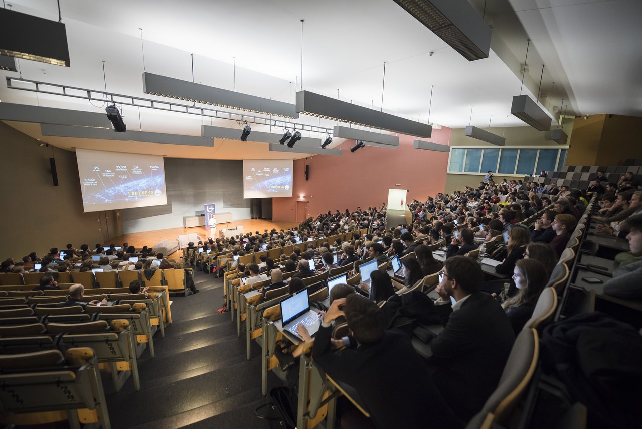 A large audience in a university lecture hall listens to a presentation with slides projected on two screens.