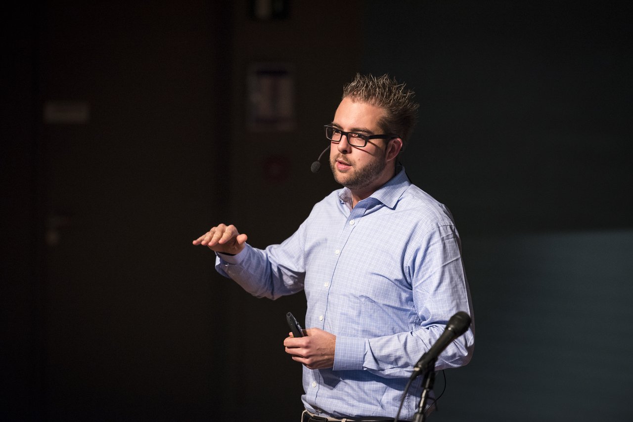 A man in a blue shirt and glasses gives a presentation, gesturing with one hand and holding a remote control.