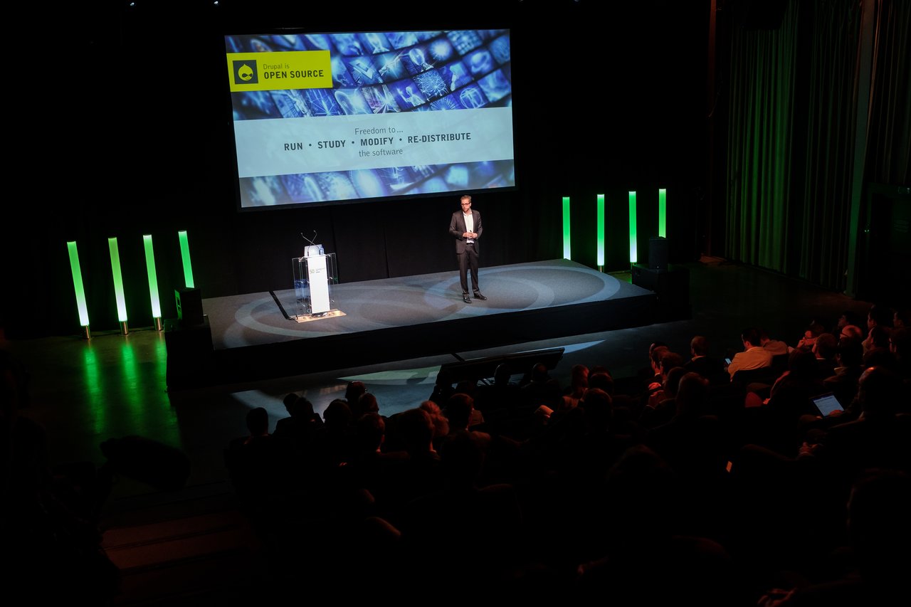 A speaker presents on stage about open-source software to an audience in a dimly lit conference hall.