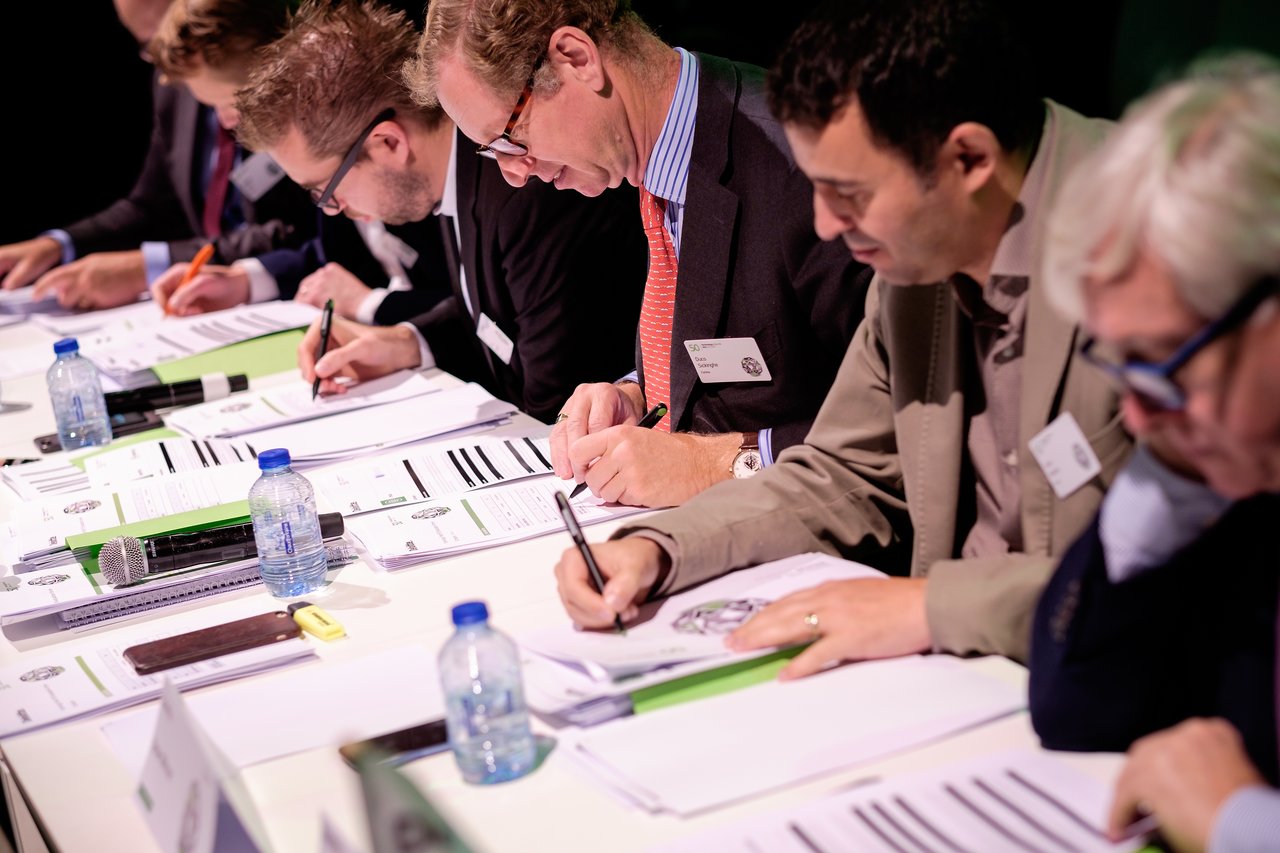 A group of professionals seated at a table, reviewing and writing on documents during a formal event or meeting.