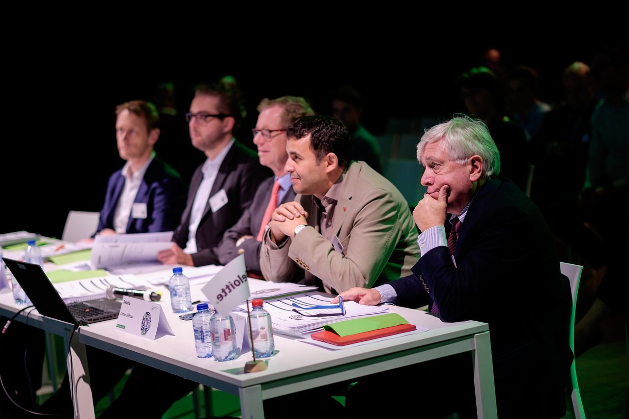 A panel of five judges sits at a table, attentively listening and reviewing documents during a professional event.
