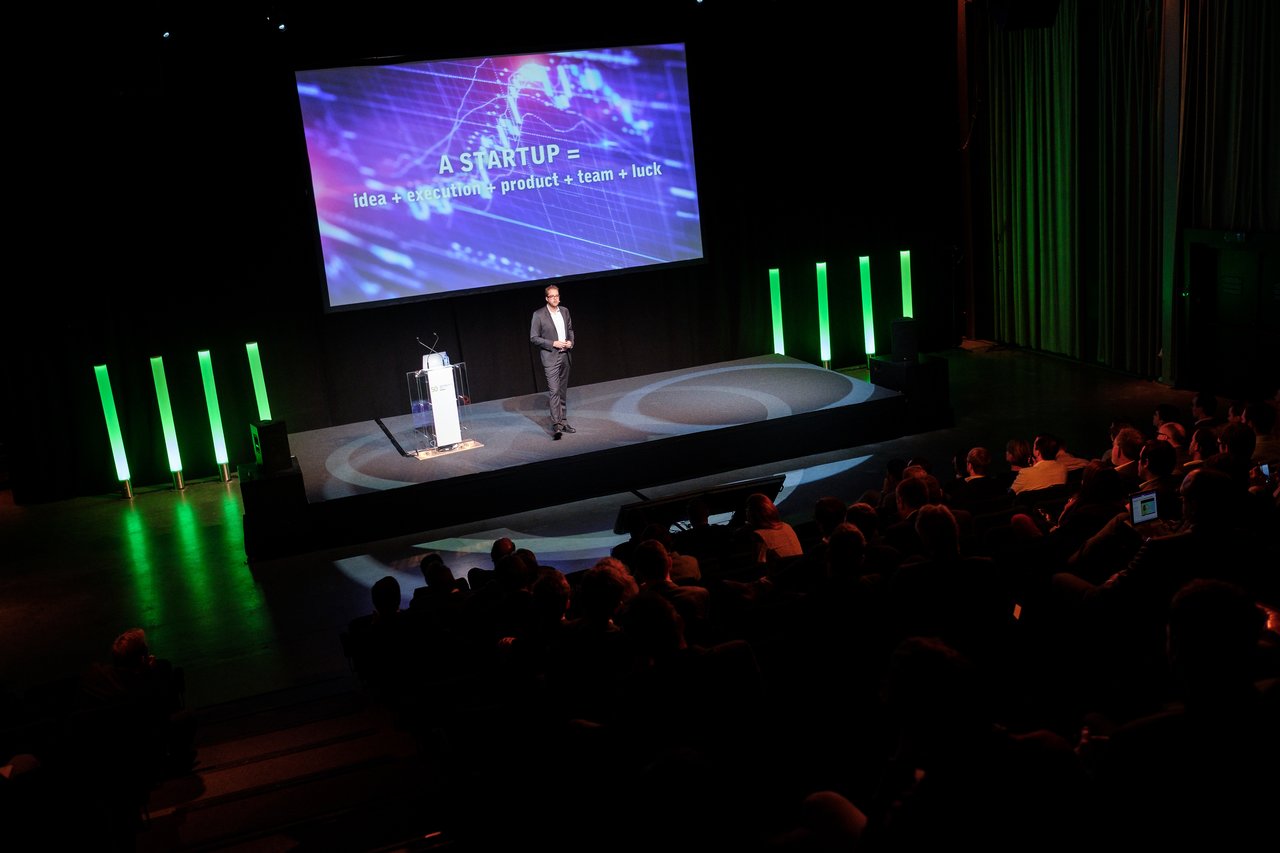 A speaker presents on stage at a conference, with a large screen displaying a startup-related slide.