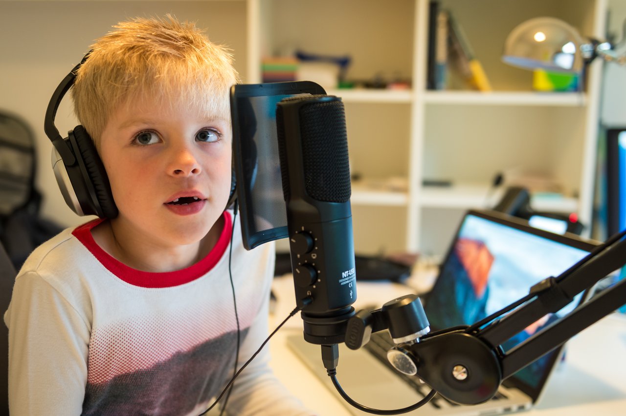 A young child wearing headphones speaks into a professional microphone in front of a computer in a home studio.
