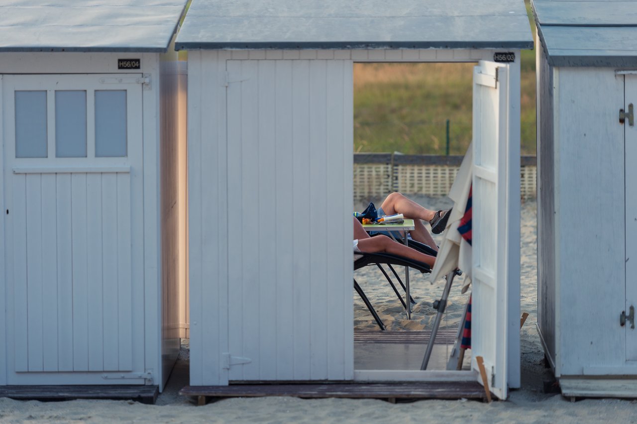 A person relaxes on a lounge chair inside a beach hut, reading a book with legs stretched out.