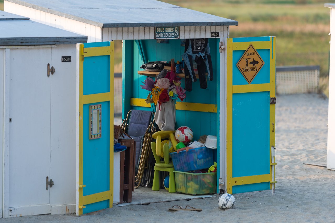 A small beach hut with open doors revealing stored beach toys, chairs, and life jackets on a sandy surface.