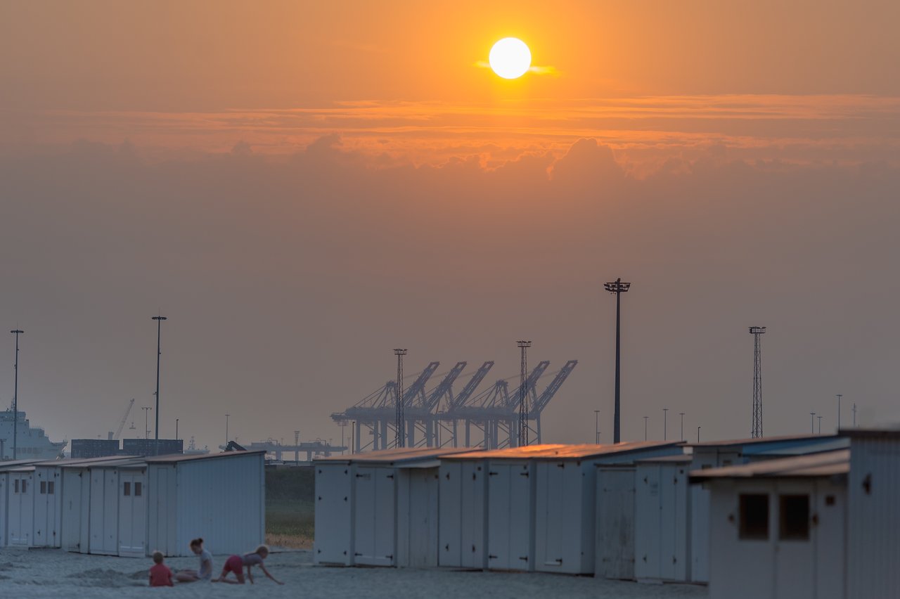 Children play on the sandy ground near beach huts, with the sun setting over industrial cranes in the background.