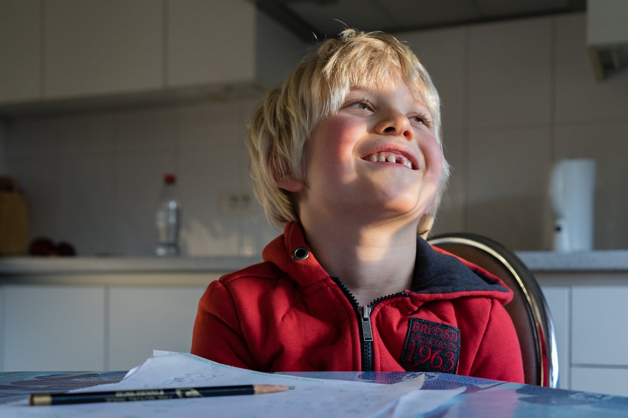 A smiling child in a red jacket sits at a table with a pencil and papers in front.