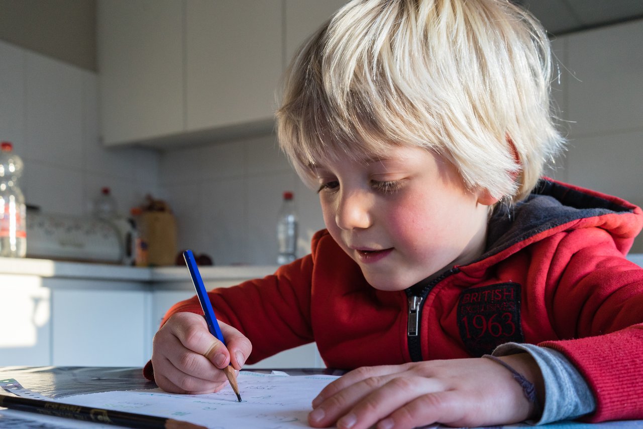 A young child in a red hoodie is drawing with a blue pencil on paper at a table.