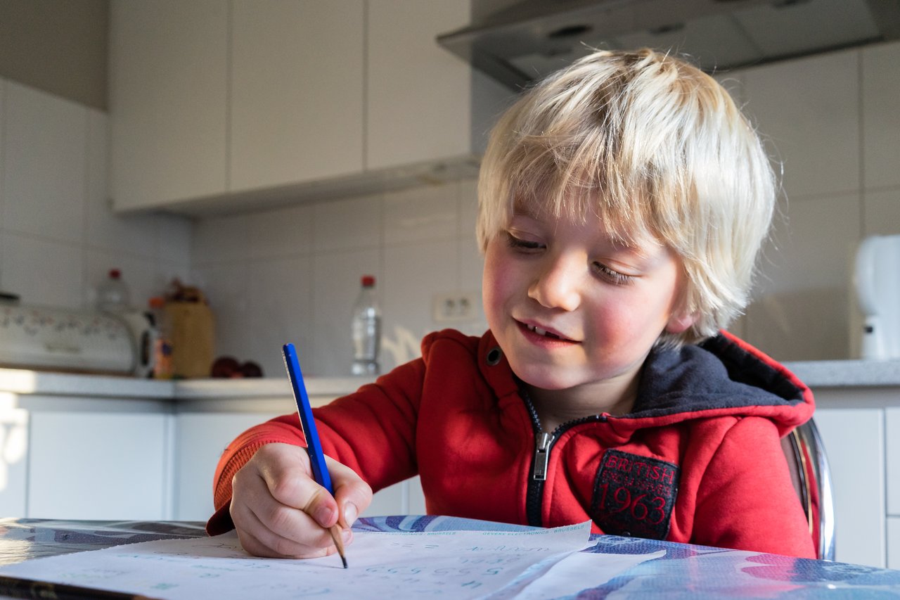 A young child in a red hoodie sits at a table, holding a blue pen and writing on paper.