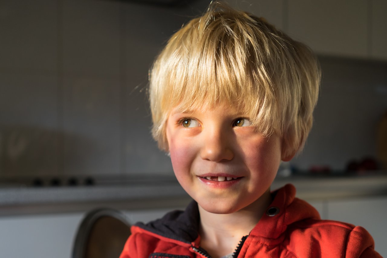 A young child with blonde hair and a red jacket smiles while looking to the side in warm lighting.