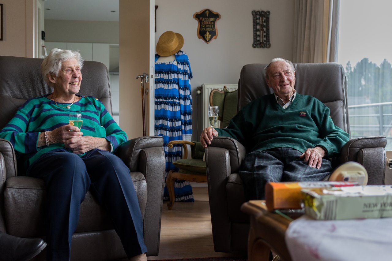 An elderly woman and man sit in armchairs, smiling and holding drinks in a cozy living room.