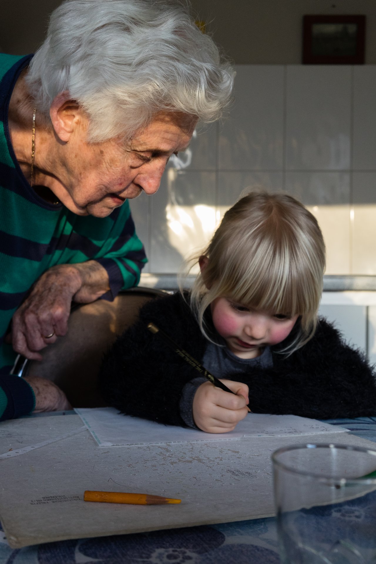 An elderly woman watches as a young child concentrates on drawing with a pencil at a table.