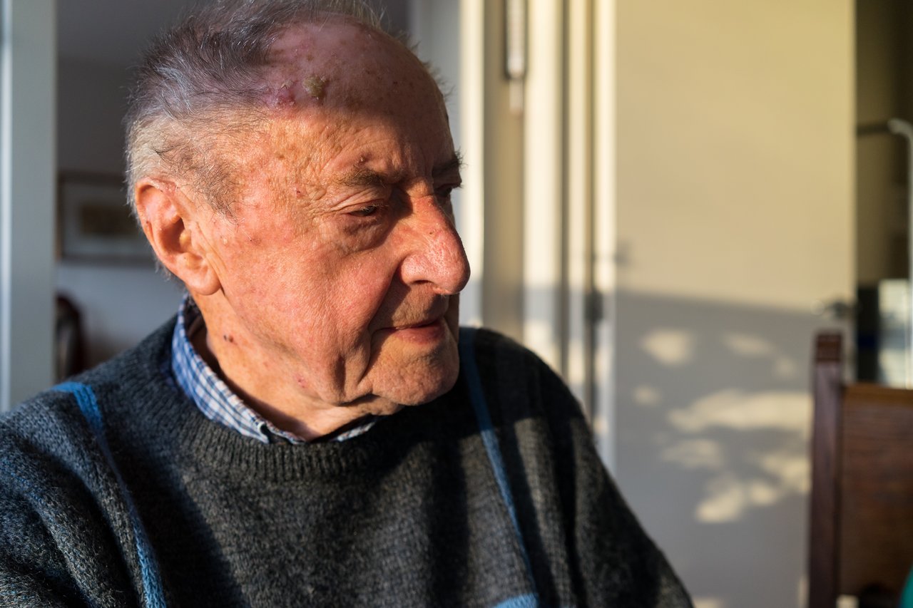 An elderly man sits in sunlight, looking to the side with a calm expression.