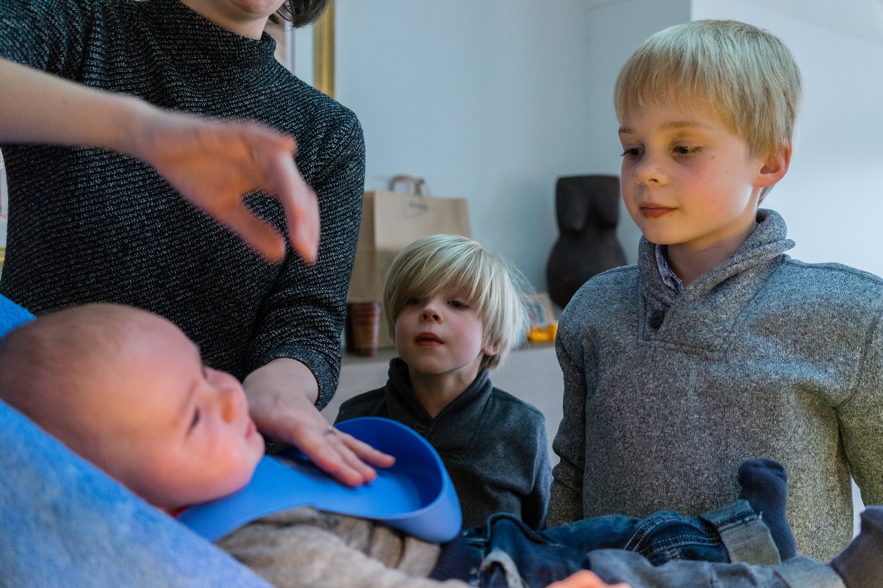 A baby is being fed with a bib on, while two young children watch closely.
