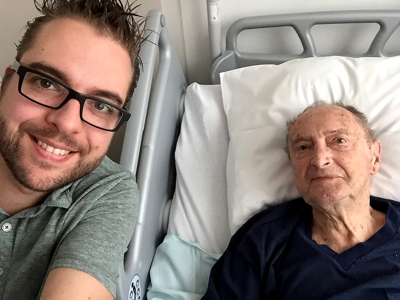 A man takes a selfie with his elderly grandfather, who is lying in a hospital bed and smiling.