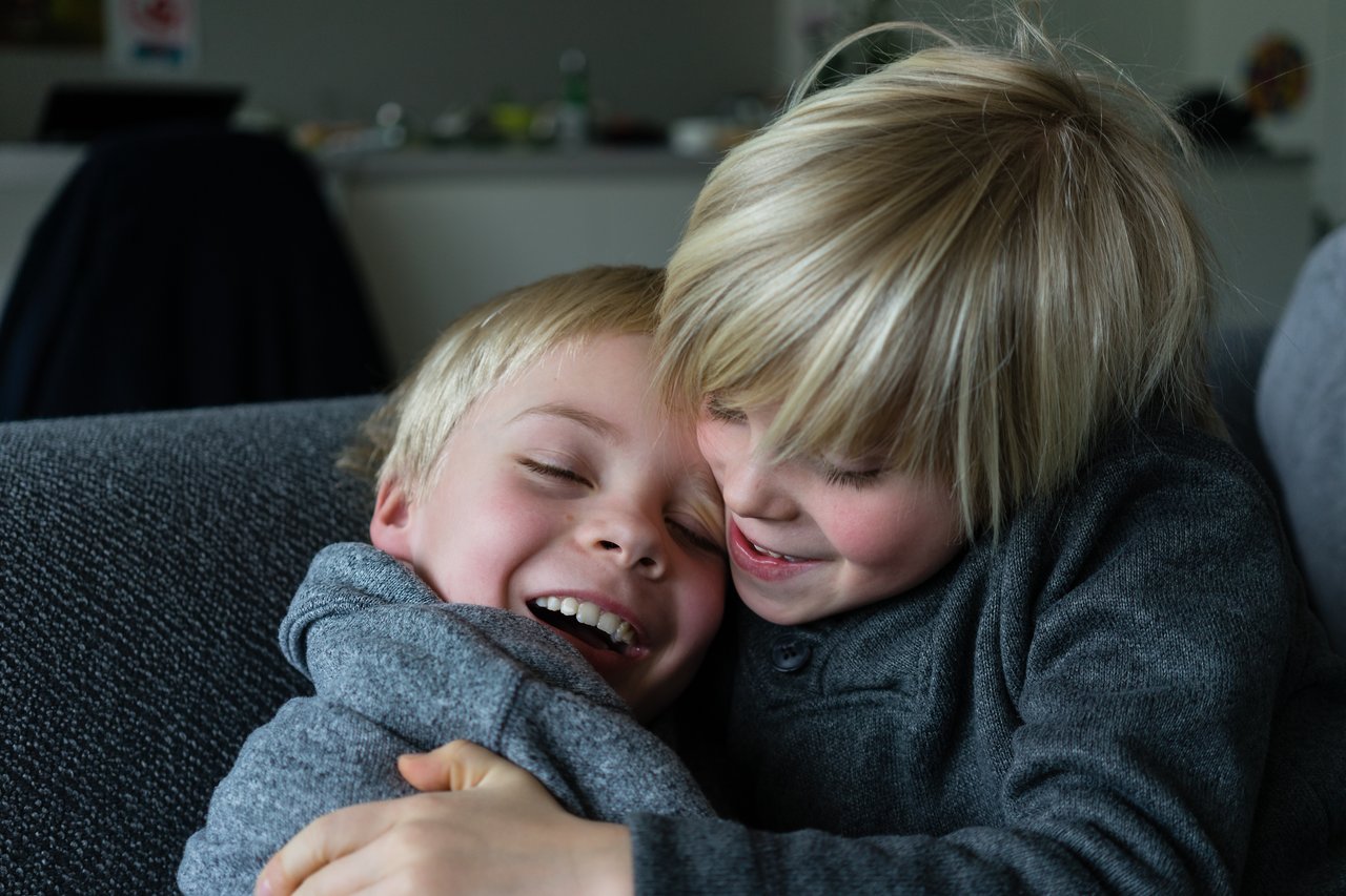 Two young brothers in matching gray sweaters share a joyful hug, smiling and laughing on a couch.