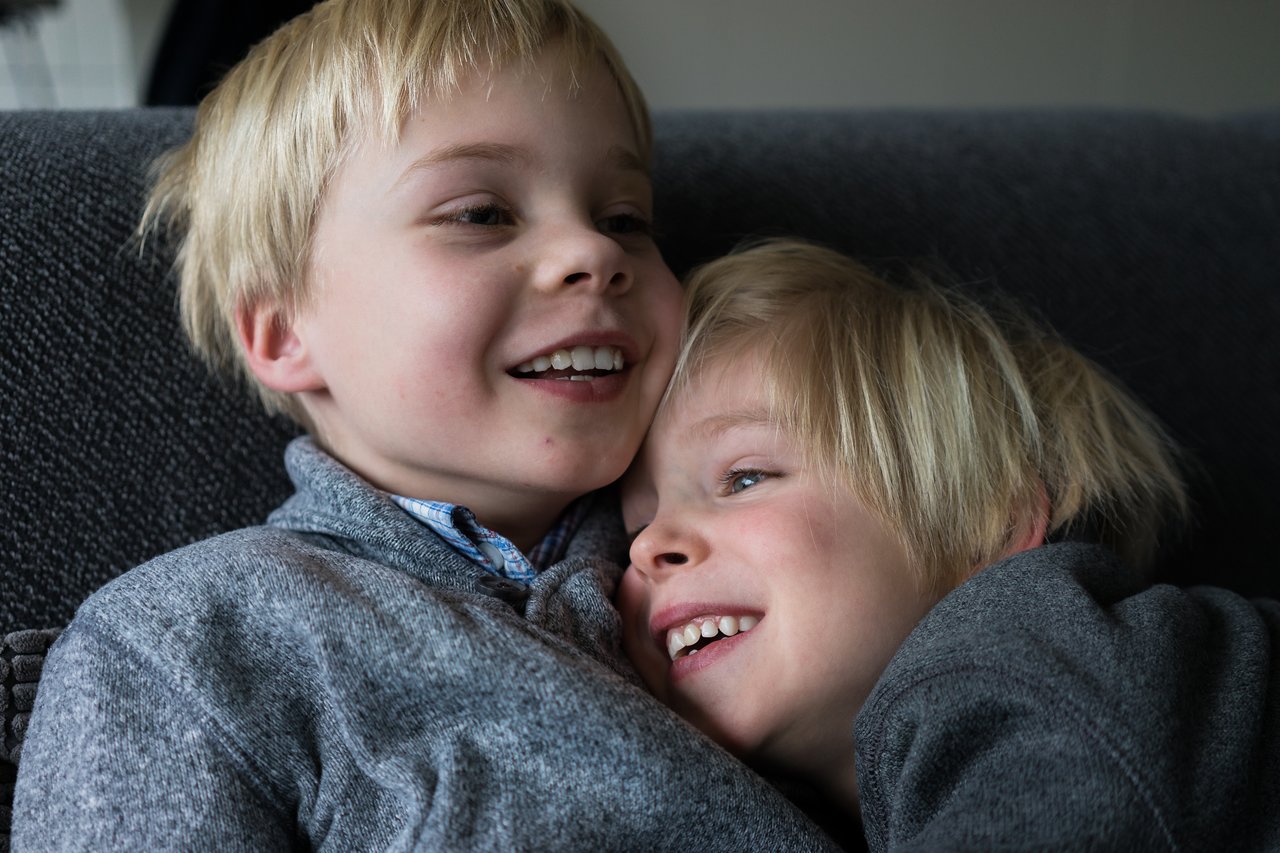 Two young boys with blonde hair and gray sweaters cuddle on a couch, smiling and laughing together.