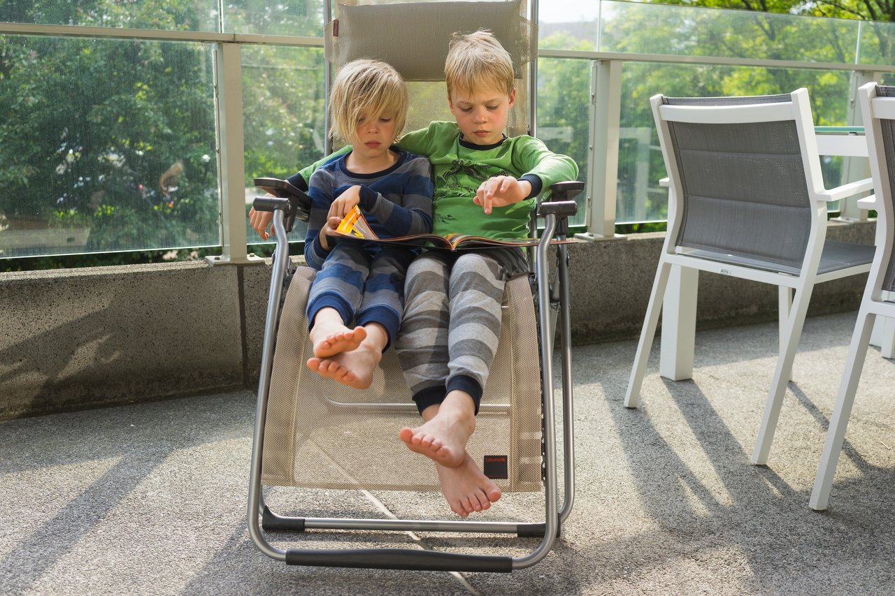 Two children sit together on a reclining chair, reading a book.