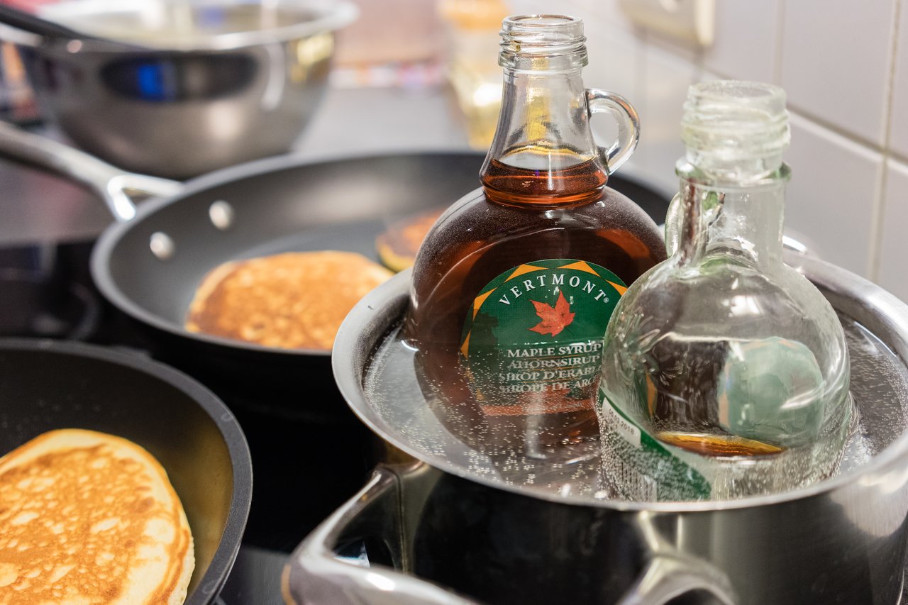 Bottles of maple syrup warming in hot water, with pancakes cooking in frying pans on the stove.