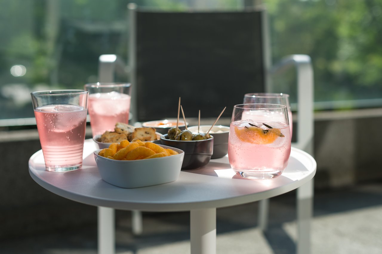 A small table with pink drinks, chips, olives, and snacks, set outdoors in the sunlight.