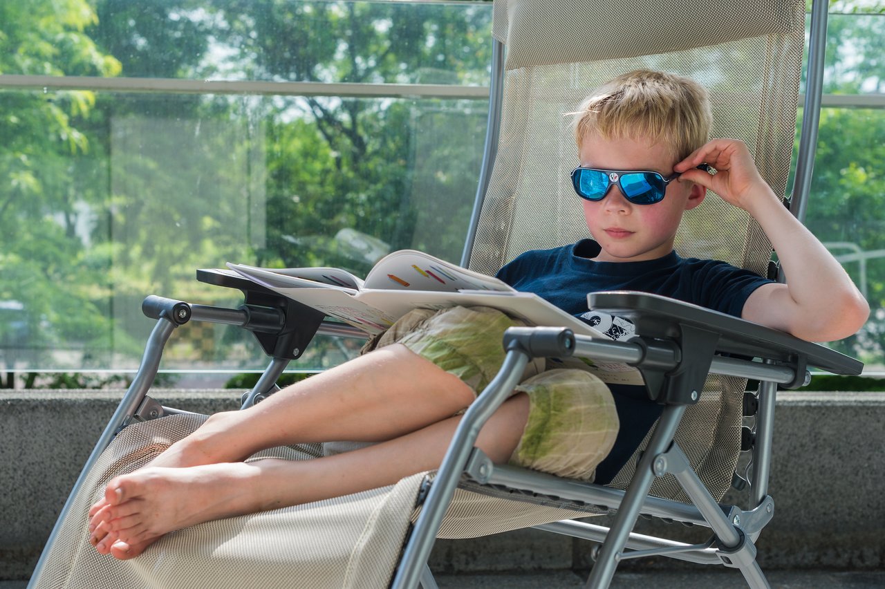A child wearing sunglasses reclines on a deck chair, reading a book with legs stretched out.