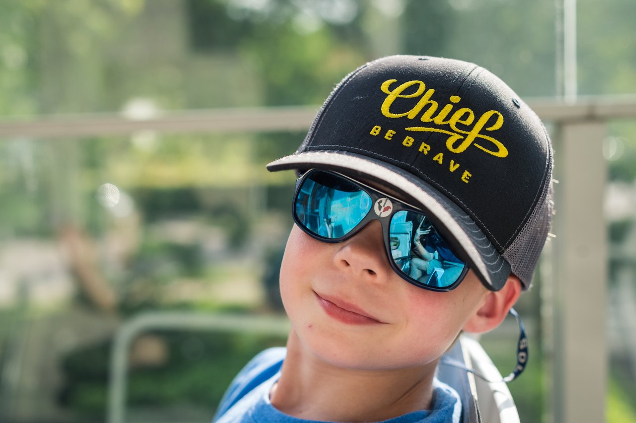 A child wearing a cap and sunglasses sits on a deck, looking relaxed.
