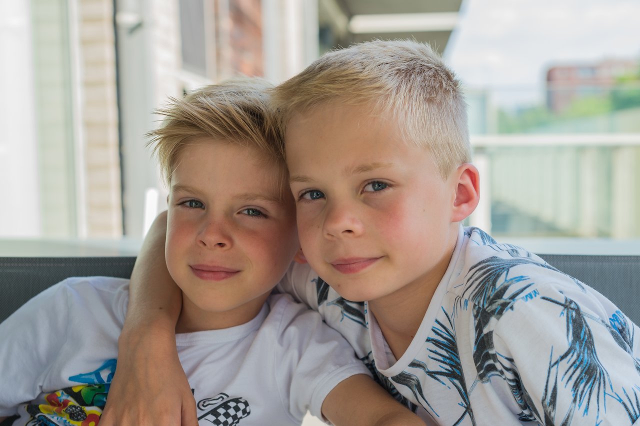 Two young boys sit closely together, one with his arm around the other, both smiling at the camera.