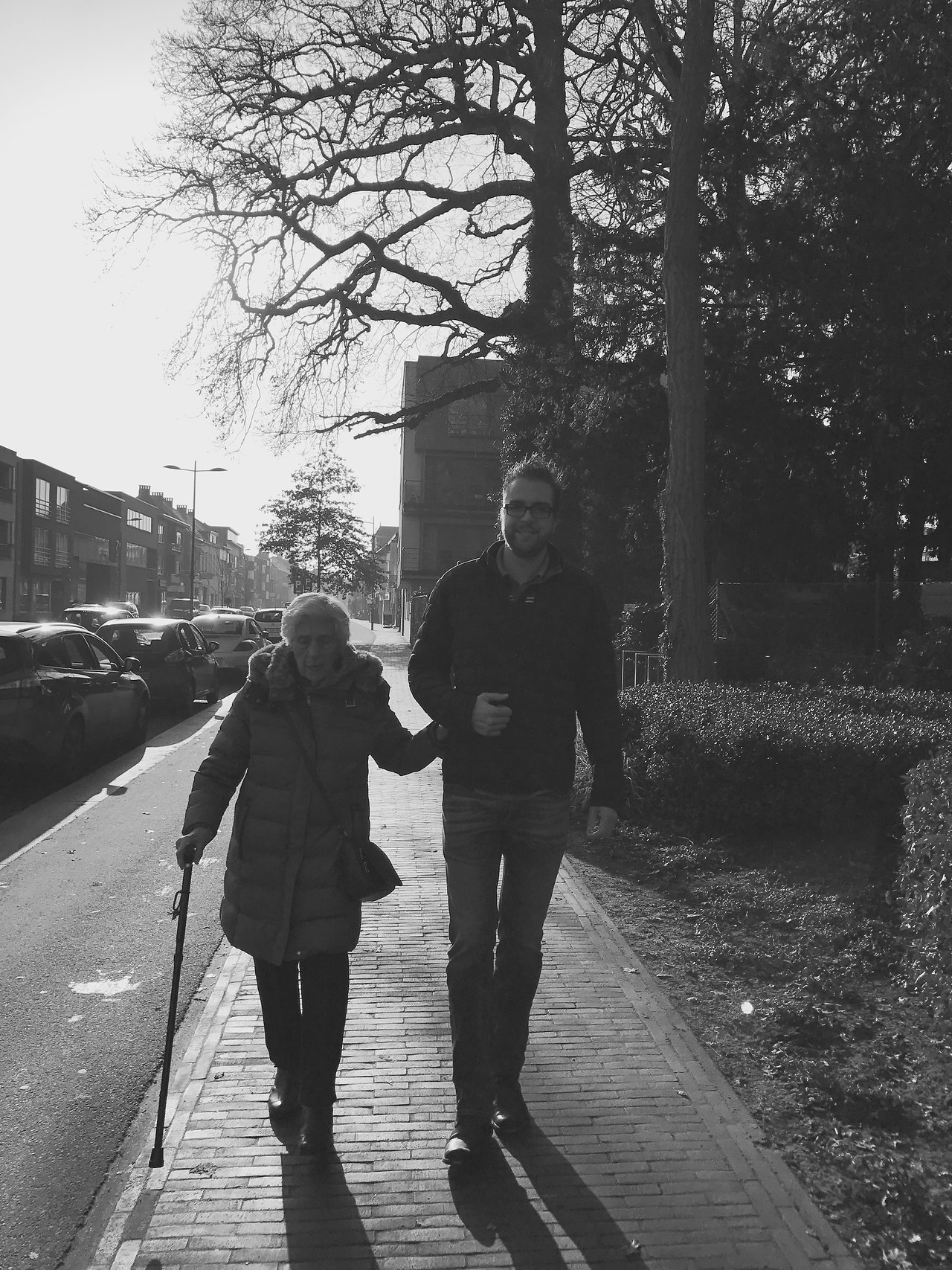 A man walks arm-in-arm with an elderly woman using a cane on a sidewalk.