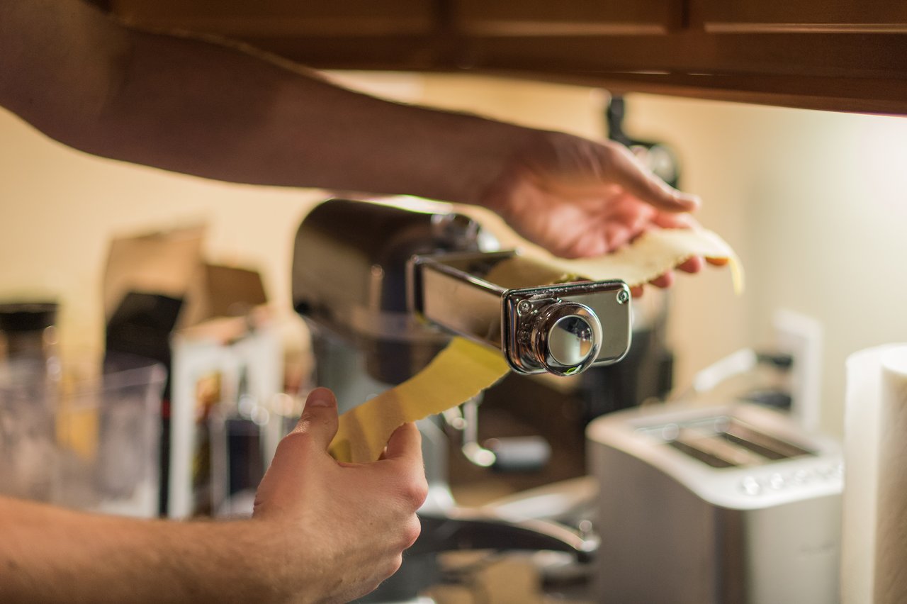 A person feeds pasta dough through a pasta machine in a kitchen, preparing homemade butternut squash ravioli.
