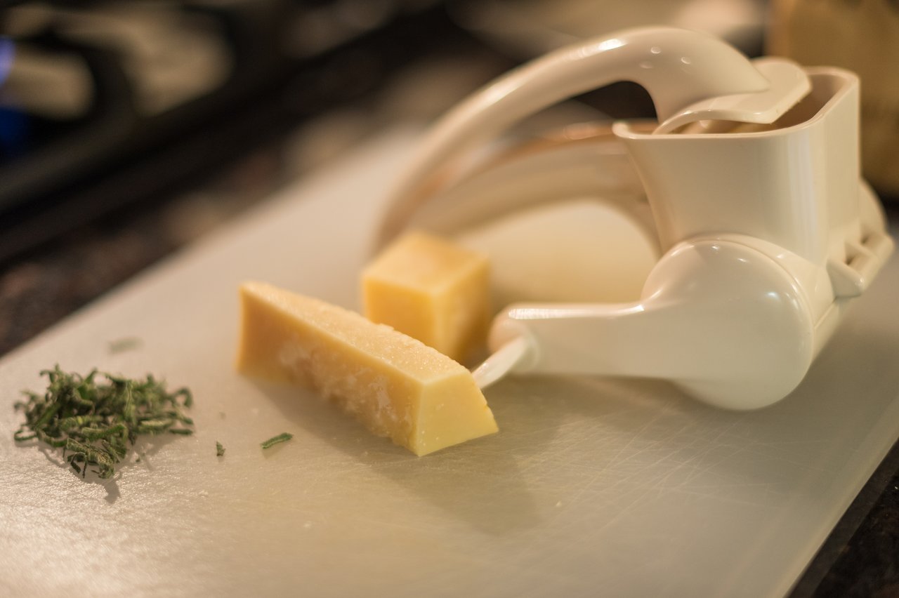A cheese grater, parmesan cheese, and chopped herbs on a cutting board, prepared for cooking.