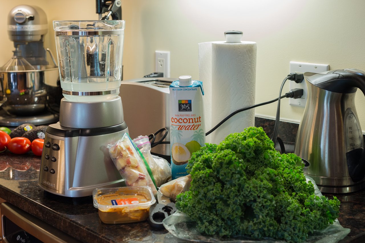 A blender with coconut water, kale, and frozen fruit on a kitchen counter, prepared for making a smoothie.
