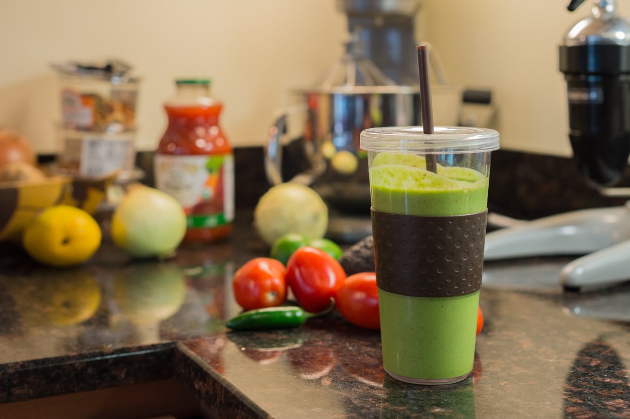 A green kale smoothie in a plastic cup with a straw, placed on a kitchen counter with vegetables.