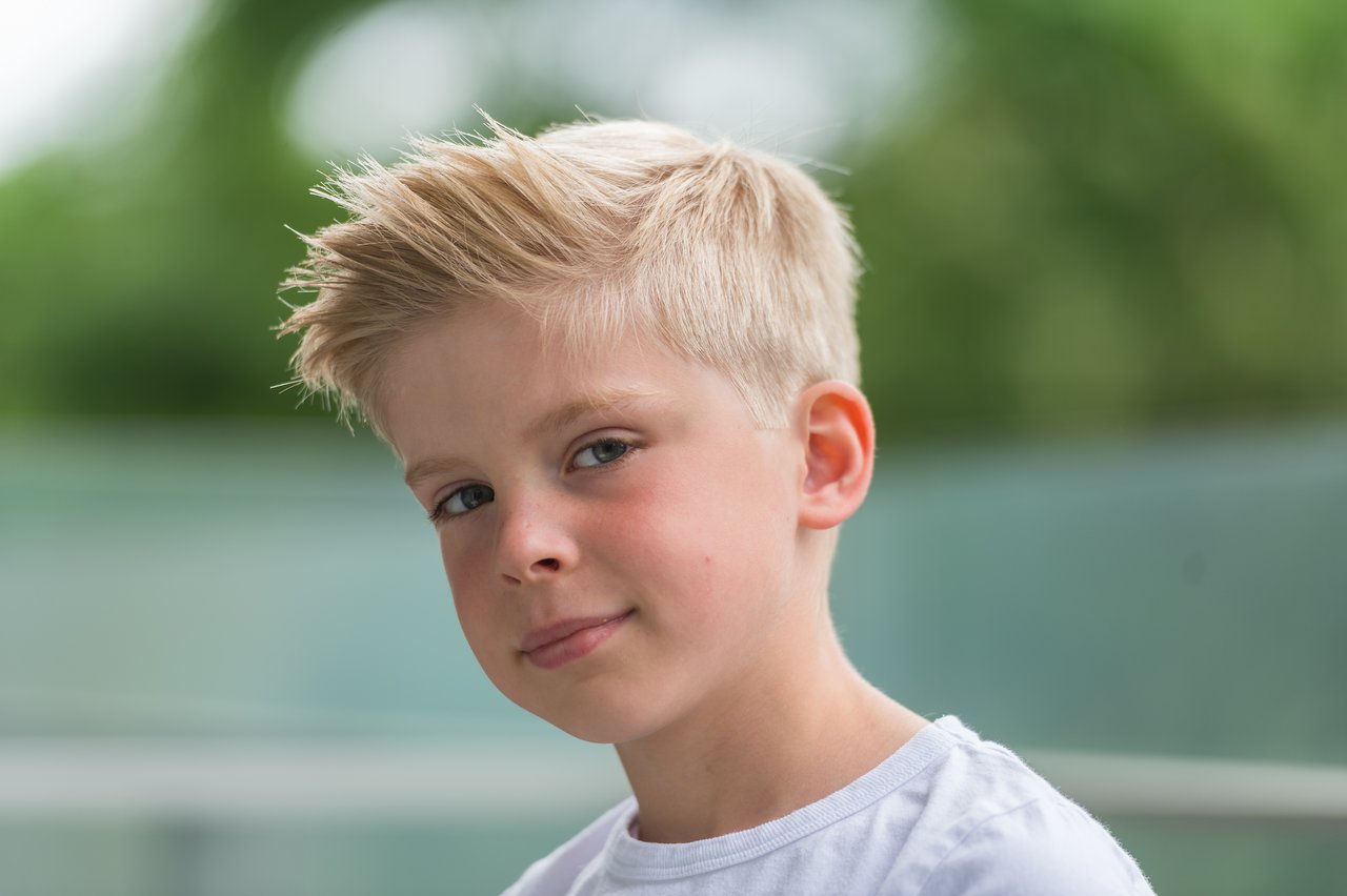 A young boy with short blond hair and a white shirt looks slightly to the side with a neutral expression.