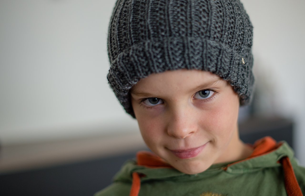 A child wearing a gray knitted hat and a green hoodie looks directly at the camera with a slight smile.
