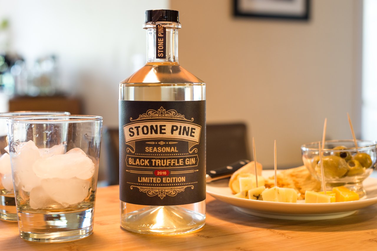 A bottle of Stone Pine Black Truffle Gin on a table with glasses of ice, cheese, and olives.