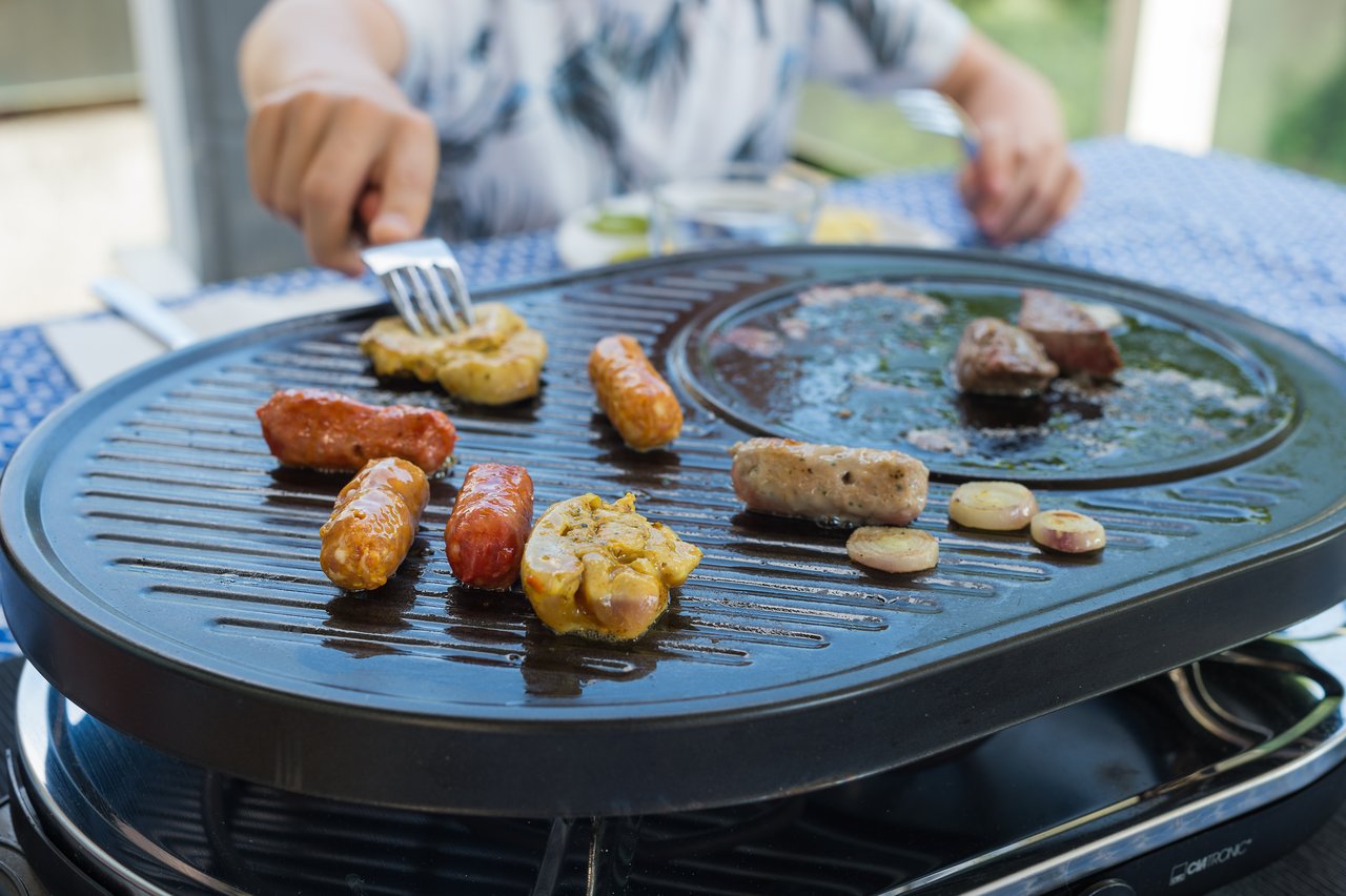 A person grills sausages, meat, and vegetables on a tabletop grill while using a fork to pick up food.