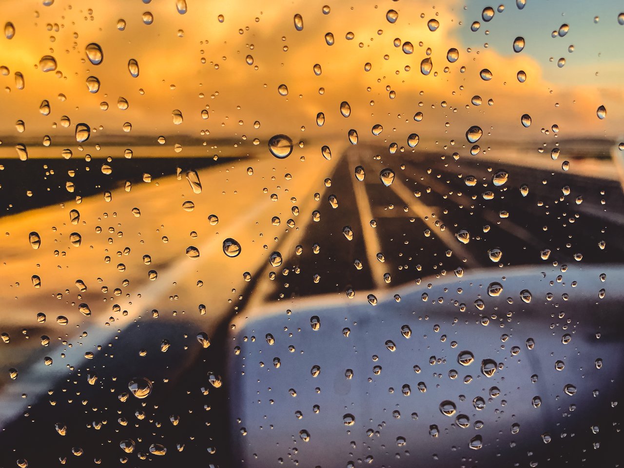 Raindrops on an airplane window with a blurred view of the wing and a colorful sky in the background.