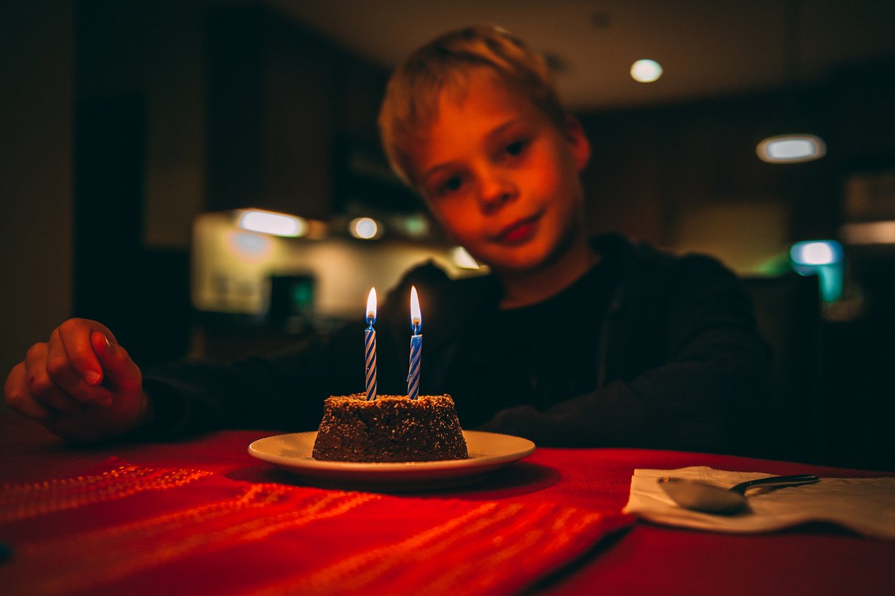 A child sits at a table, looking at a small birthday cake with two lit candles.