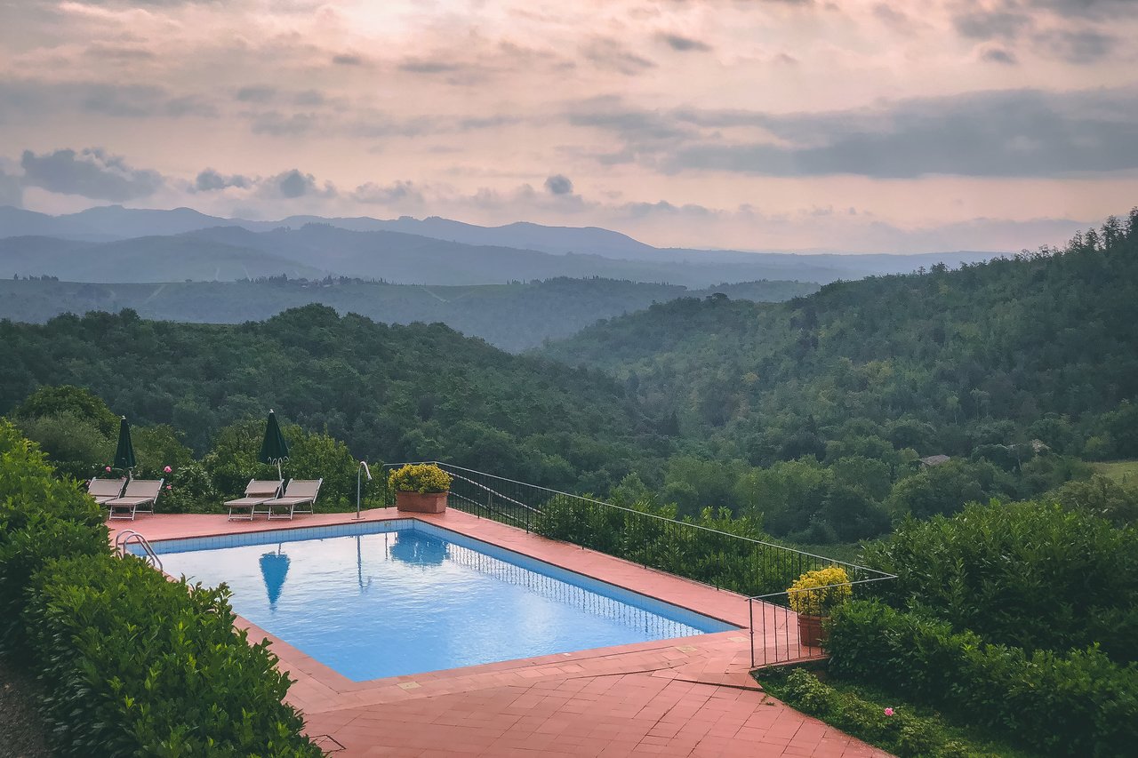 A swimming pool with lounge chairs and umbrellas overlooks green hills and distant mountains under a cloudy sky.