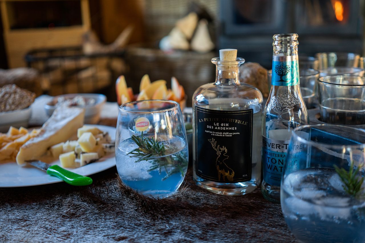 A bottle of "Le Gin des Ardennes" with a gin and tonic garnished with rosemary on a table.