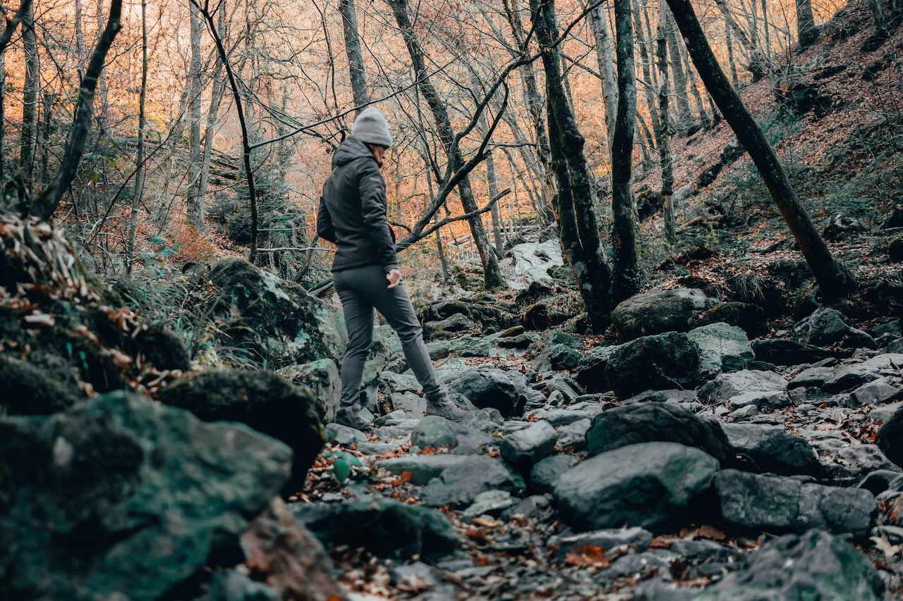 A person wearing a jacket and beanie walks on a rocky forest trail surrounded by trees with autumn leaves.