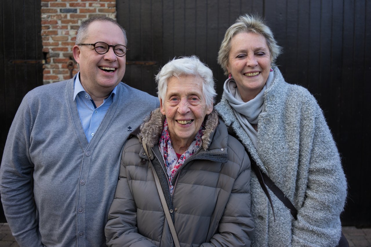 An elderly woman smiles between a man and a woman, all dressed warmly, celebrating her ninetieth birthday.