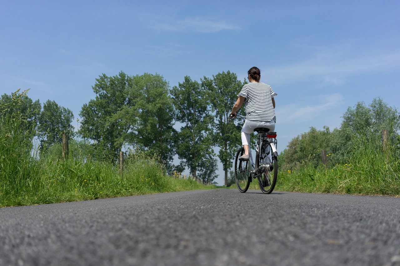 A person wearing a striped shirt and white pants rides a bicycle on a rural paved road surrounded by greenery.