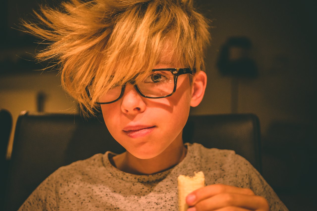 A young person with messy blond hair and glasses sits indoors, holding a piece of food and looking at the camera.