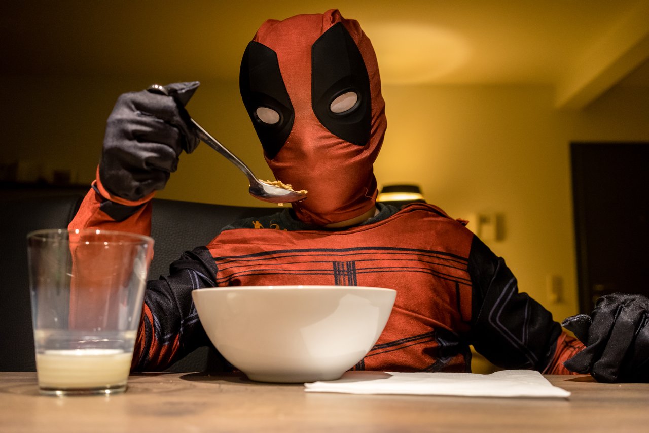 A person dressed as Deadpool sits at a table, eating cereal with a spoon.