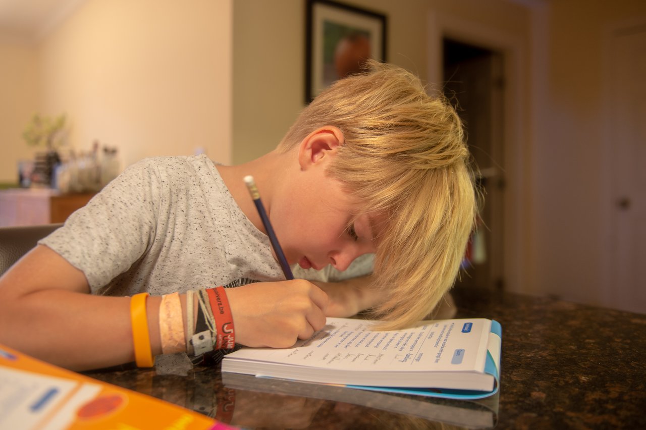 A boy is sitting at a table, writing in a workbook with a pencil.