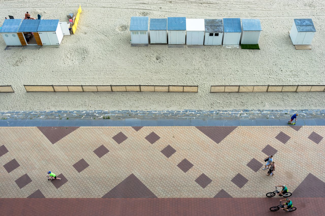 People walk, cycle, and skateboard on a patterned promenade near a sandy beach with small beach huts.