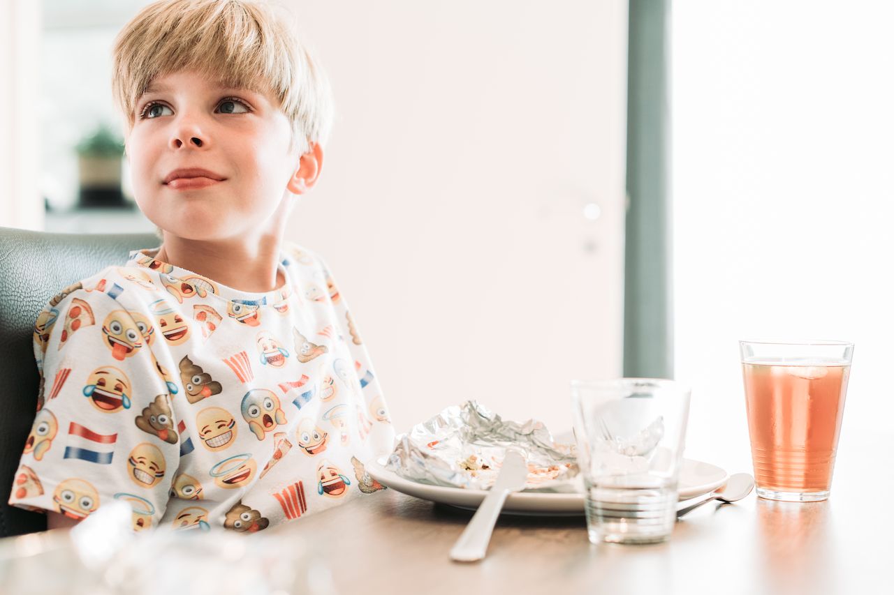 A child sits at a table with a plate of food and a drink, looking off to the side.