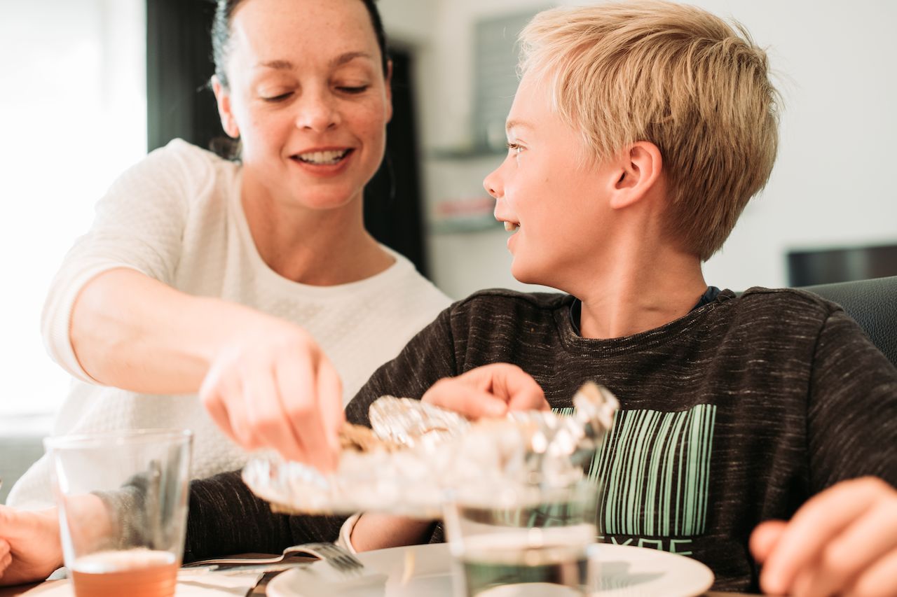 A woman and a boy sit at a table, smiling as she serves food from a foil dish.